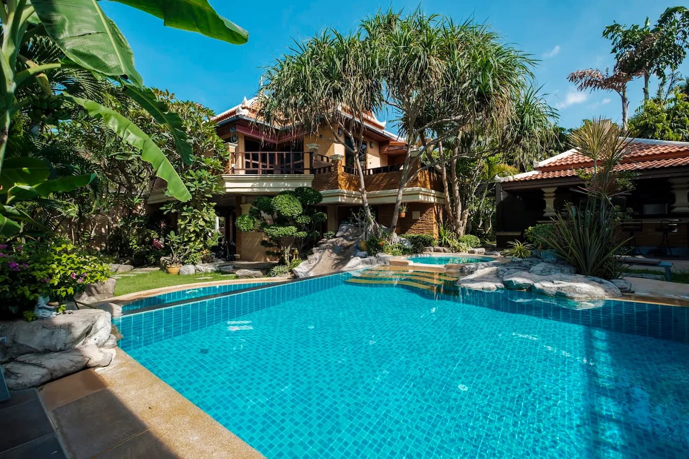Tropical villa with a blue tiled pool, stone slide, and lush greenery under clear skies.