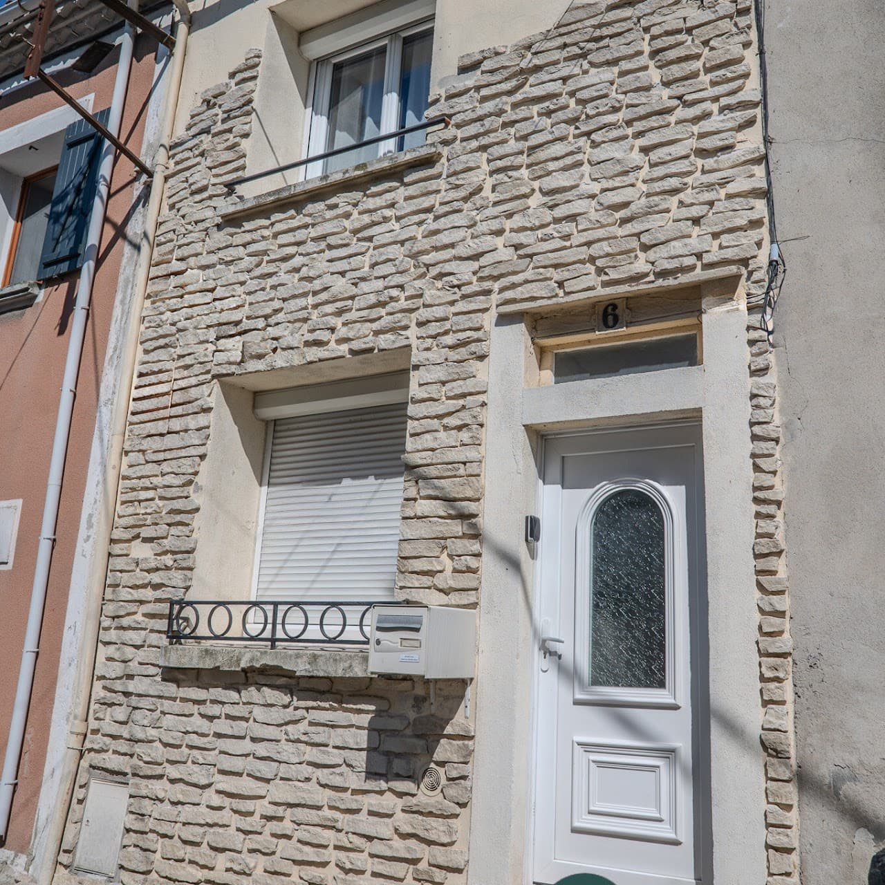 Narrow stone-clad house facade with a white door, shuttered window, and a white mailbox.