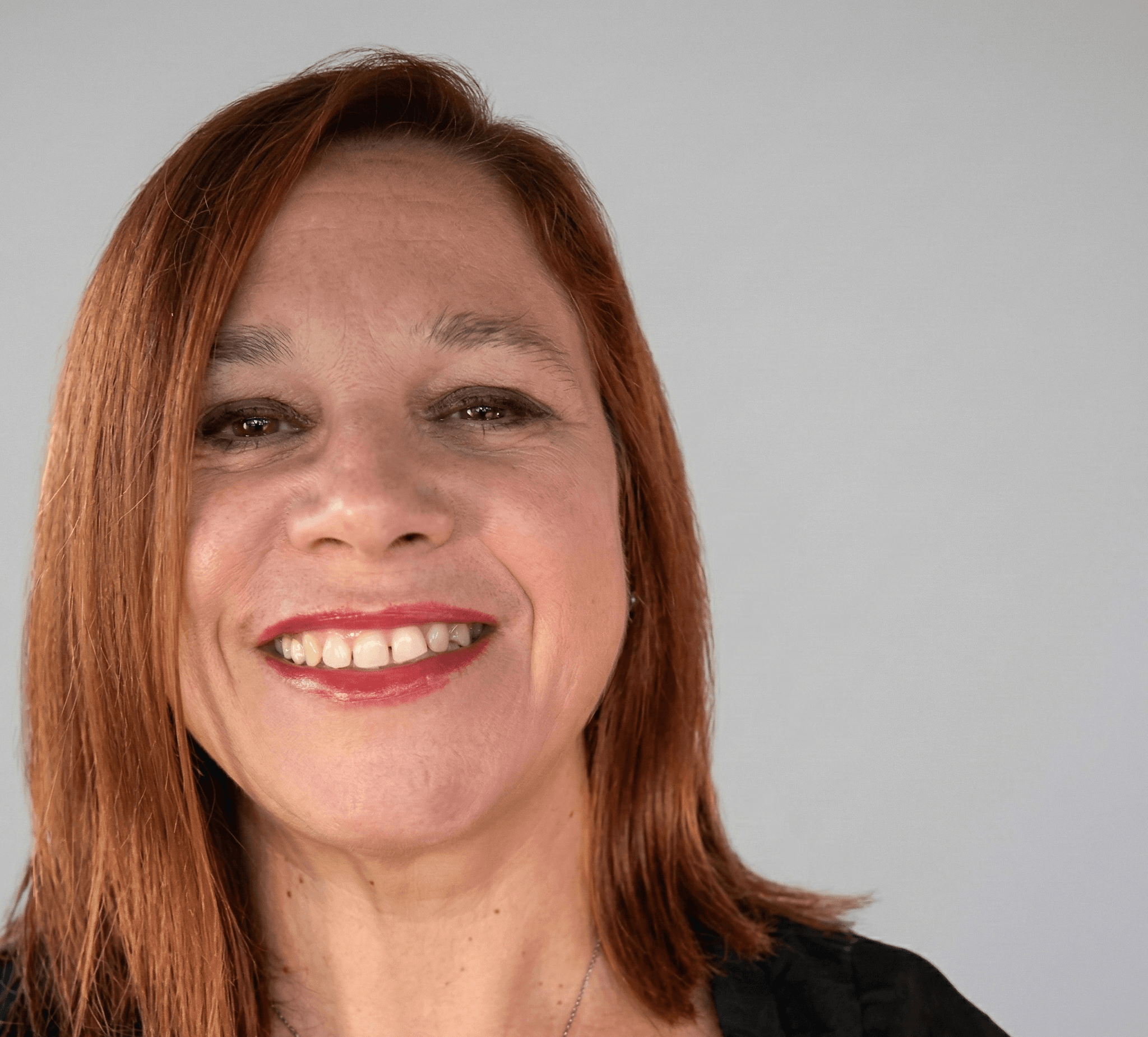 Une femme souriante aux cheveux roux et rouge à lèvres rouge sur fond gris.